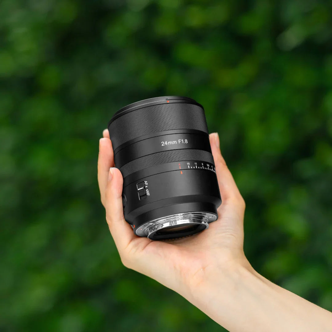 Hand holding a 24mm F1.8 camera lens with autofocus and manual focus switch on green blurred background