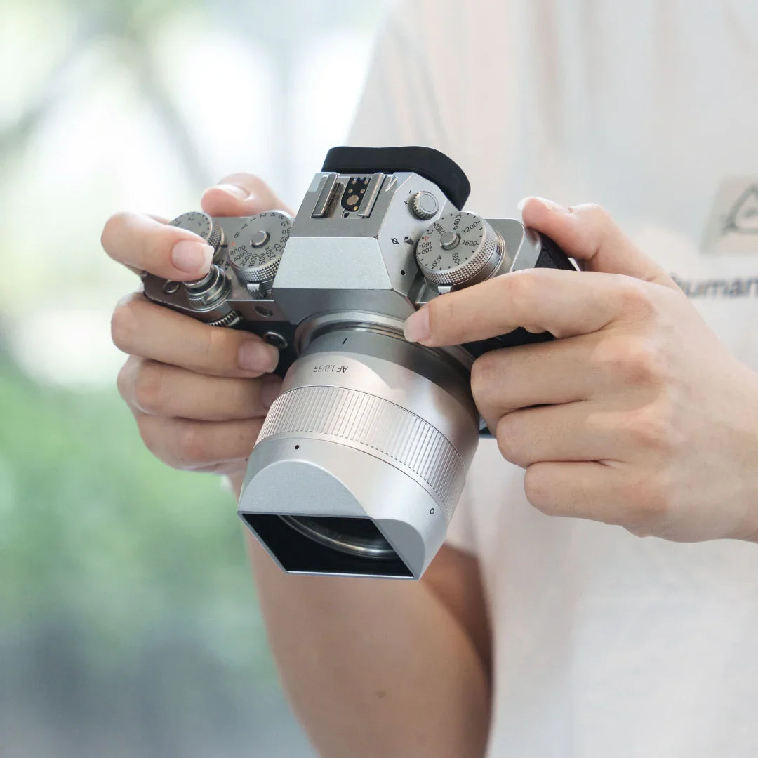 Person holding a silver retro-style digital camera with manual dials and a large lens indoors