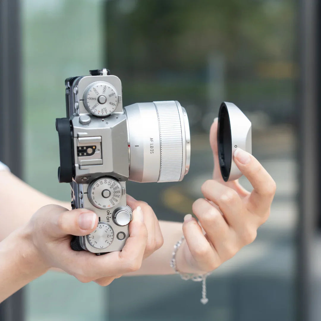 Person holding a silver vintage-style camera with manual dials and removing the lens cap outdoors