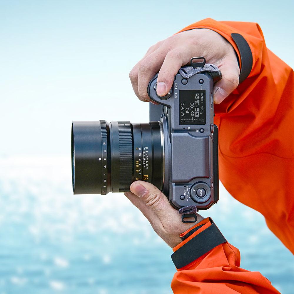Hands holding a camera with 90mm F1.25 lens against a blurred ocean background