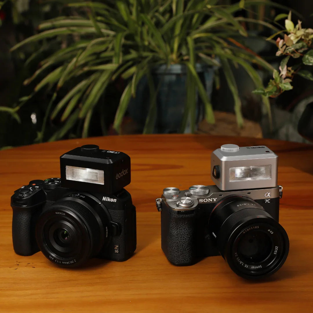 Nikon Z30 and Sony Alpha 7C cameras with mounted Godox and Nissin external flash units on wooden table